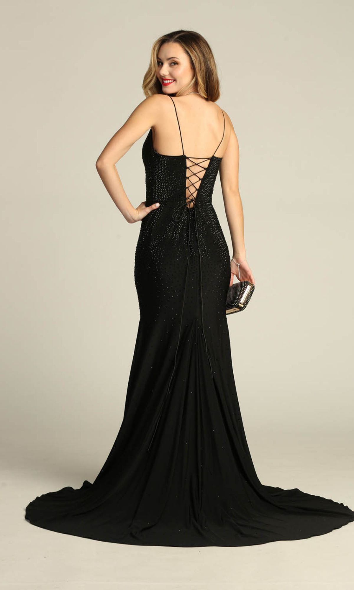 A woman in the Chicas YG5026 Lace-Up Long Beaded Prom Dress faces backward, showcasing its thin straps and lace-up detail, while holding a small clutch.
